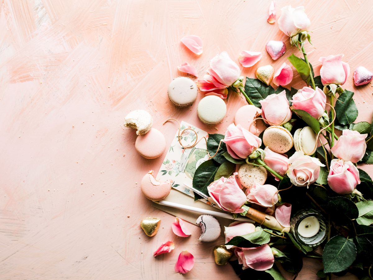 The image shows a flat lay of pink roses, macarons, small heart-shaped chocolates, and a candle on a light pink background.