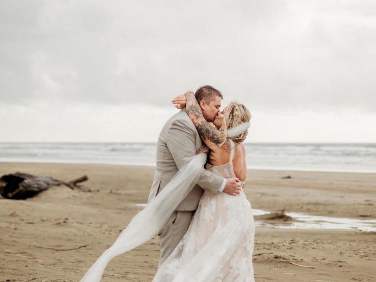 A couple in wedding attire embraces on a sandy beach, with a cloudy sky and ocean in the background.