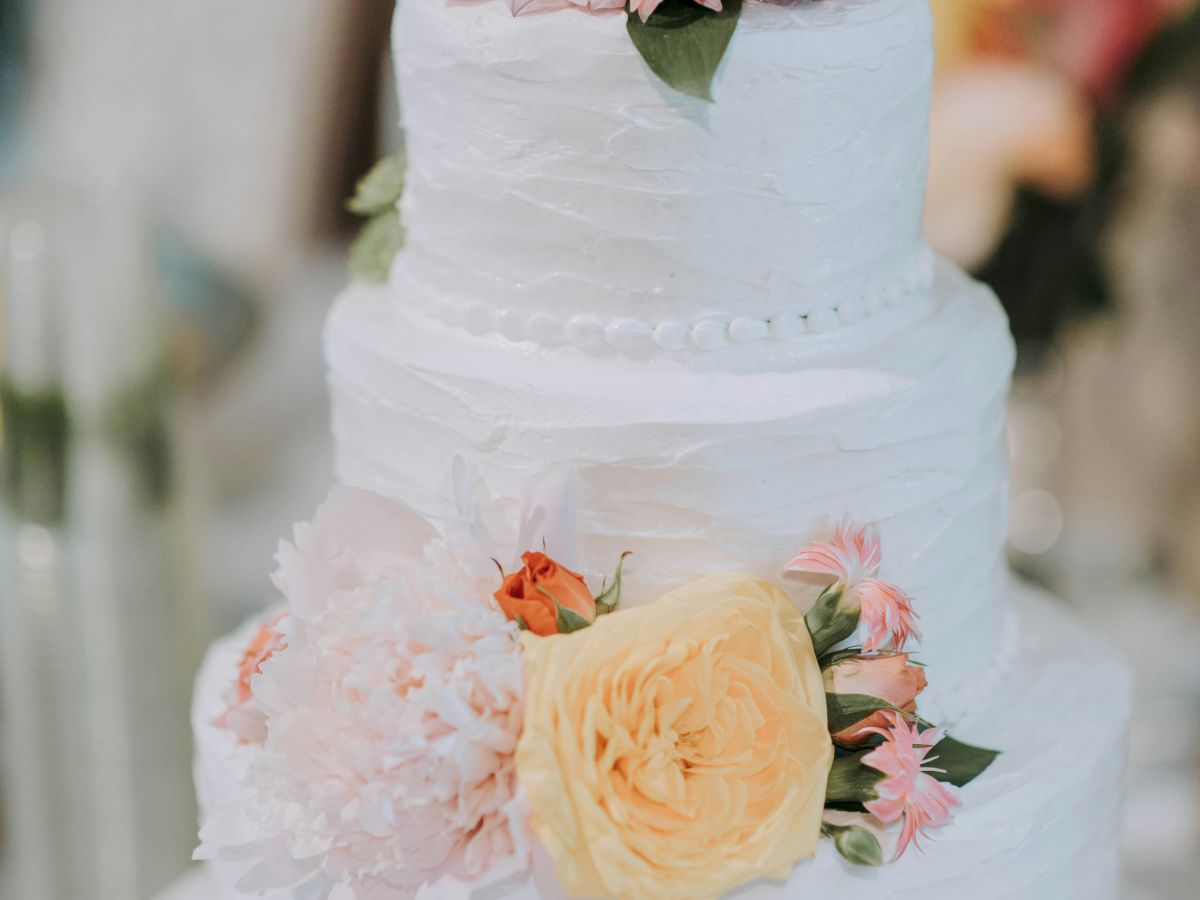 A three-tier white cake adorned with colorful flowers, including yellow and pink blossoms, displayed on a stand.