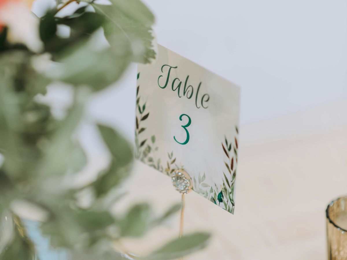 A Table 3 card with floral design is displayed on a stand surrounded by greenery and a glass votive holder.