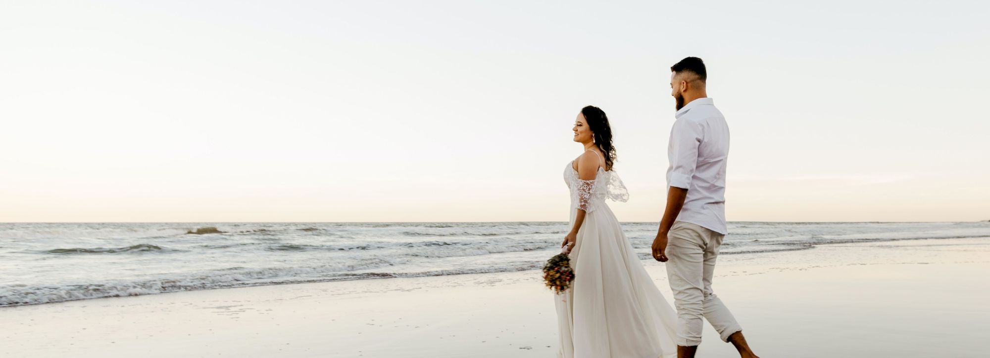 A couple walks on a beach at sunset, with the woman in a flowing white dress and the man in a white shirt and pants, enjoying the view.