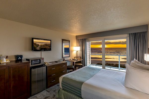 A cozy hotel room with a double bed, TV, mini-fridge, desk, and large windows showcasing a sunset view over the ocean.