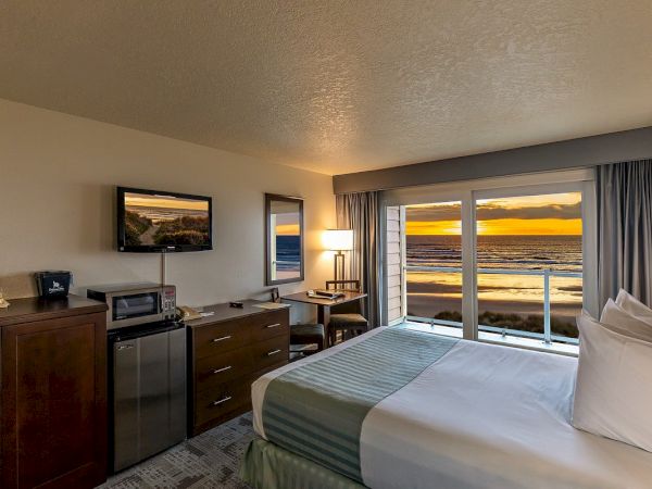 A cozy hotel room with a double bed, TV, mini-fridge, desk, and large windows showcasing a sunset view over the ocean.