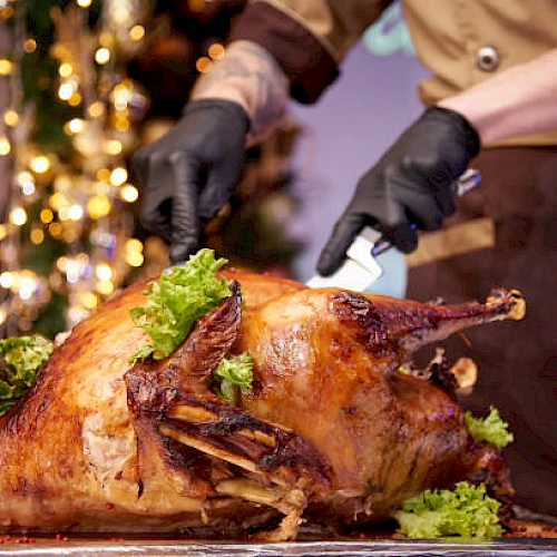 A roasted turkey being carved at the table with herbs, gloved hands, and festive lights in the background.