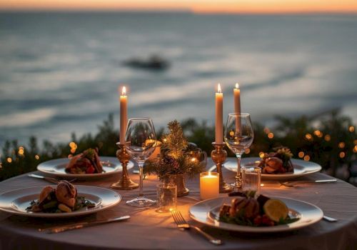 A romantic seaside dinner setup with candles, wine glasses, plates of food, and a sunset over the ocean.
