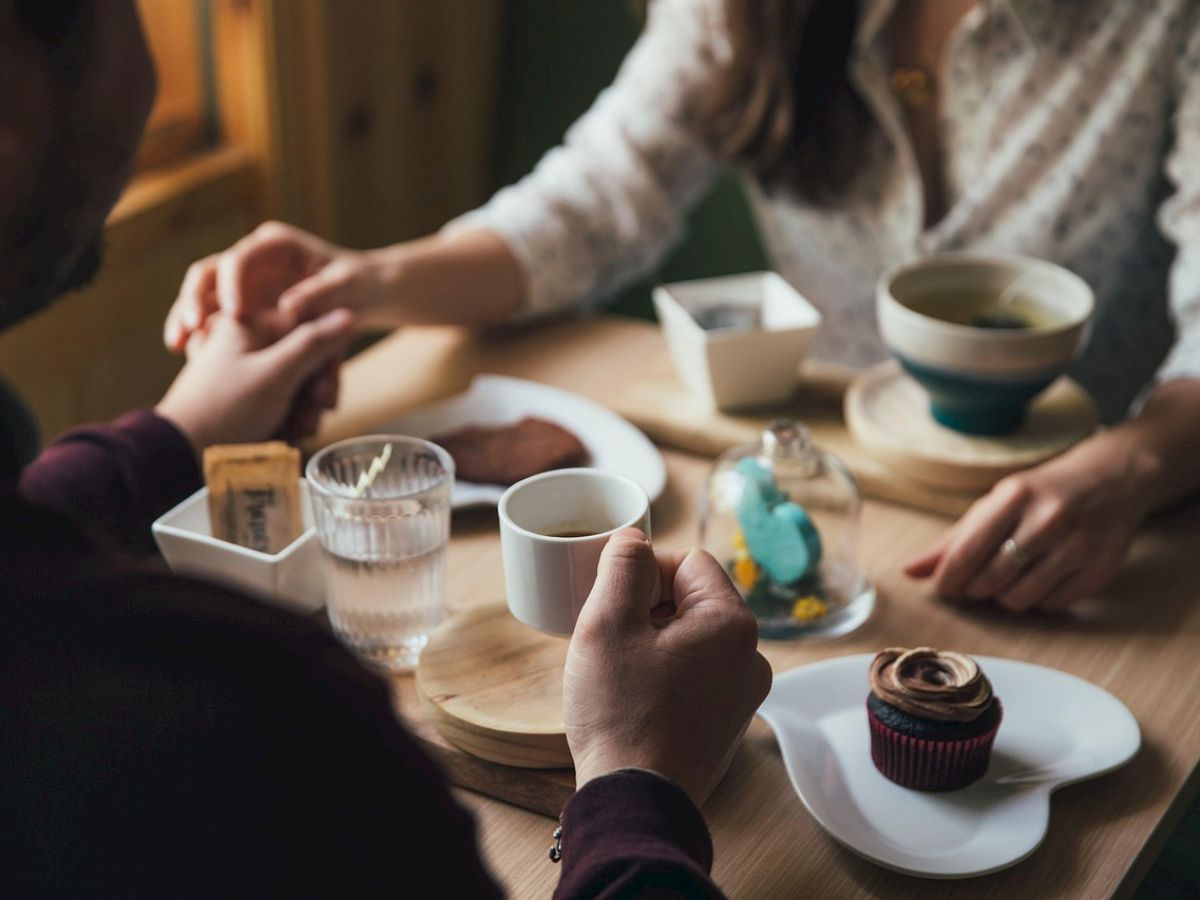 Two people sit at a wooden table with coffee cups, a glass of water, and a cupcake on a plate; they hold hands, sharing a quiet moment.