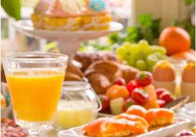 A vibrant breakfast spread with orange juice, pastries, fresh fruit, croissants, muffins, and a cake in a sunlit, festive setting.