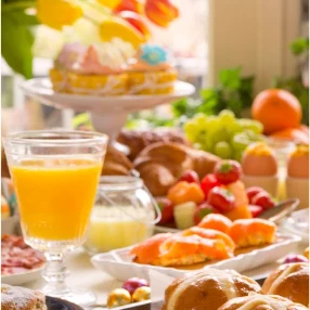 A vibrant breakfast spread with orange juice, pastries, fresh fruit, croissants, muffins, and a cake in a sunlit, festive setting.