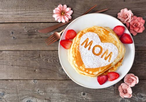 A stack of pancakes on a plate with &ldquo;MOM&rdquo; written in syrup, surrounded by strawberries and pink flowers on a wooden table.