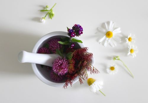 A white mortar with purple and pink flowers, plus a few stems; scattered white daisies with yellow centers on a light surface.
