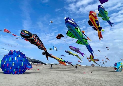 Kite festival on the beach with colorful fish-shaped kites soaring in a blue sky, people strolling, and a large blue balloon in the sand.