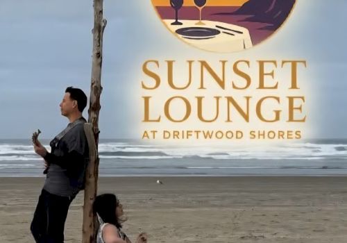 Two people on a beach near a sign that reads &ldquo;Sunset Lounge at Driftwood Shores&rdquo; with a sunset logo, the man leaning on a post and the woman seated on the sand.