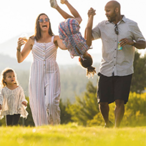 A family of four is enjoying outdoor time. The mother and father are swinging a child, while another child looks on, all smiling.