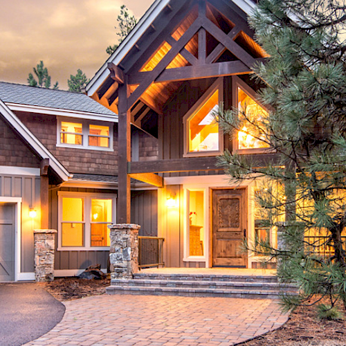 A beautiful, large house with warm lighting, wooden accents, and surrounded by trees. The design features a mixed material facade.