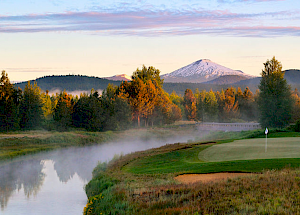 A serene landscape with a golf course beside a misty river, surrounded by trees and mountains in the distance under a clear sky with scattered clouds.