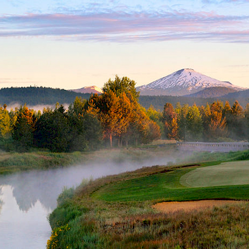 A serene landscape with a golf course beside a misty river, surrounded by trees and mountains in the distance under a clear sky with scattered clouds.