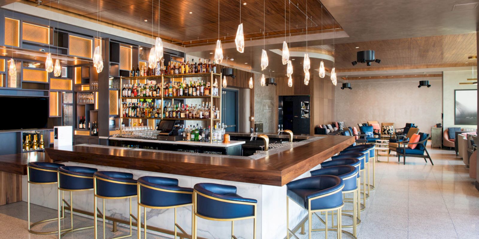The image shows a modern, well-lit bar with blue bar stools, a wooden counter, hanging lights, and various bottles on display, located in a stylish lounge area.