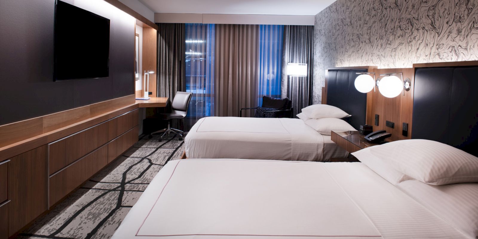 This image shows a modern hotel room with two neatly made beds, a wall-mounted TV, a desk with a chair, and contemporary decor.
