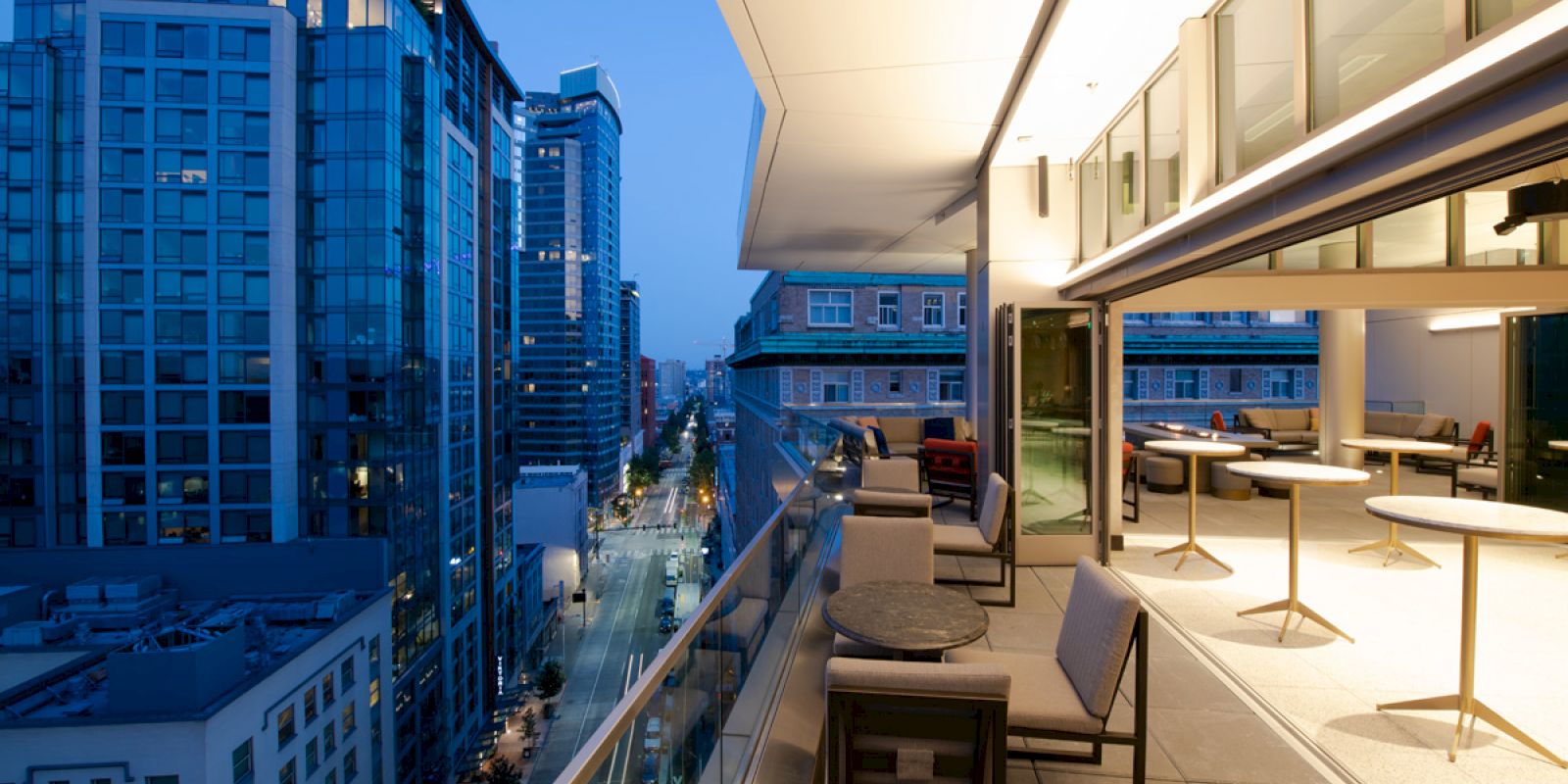 The image shows a modern outdoor balcony at dusk with furniture, overlooking a city street lined with tall buildings and illuminated windows.