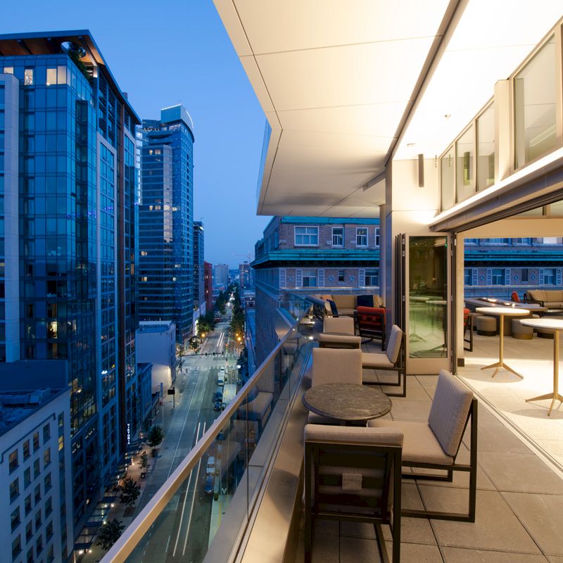 A modern cityscape with high-rise buildings at dusk, featuring a stylish balcony with tables and chairs overlooking the urban street below.