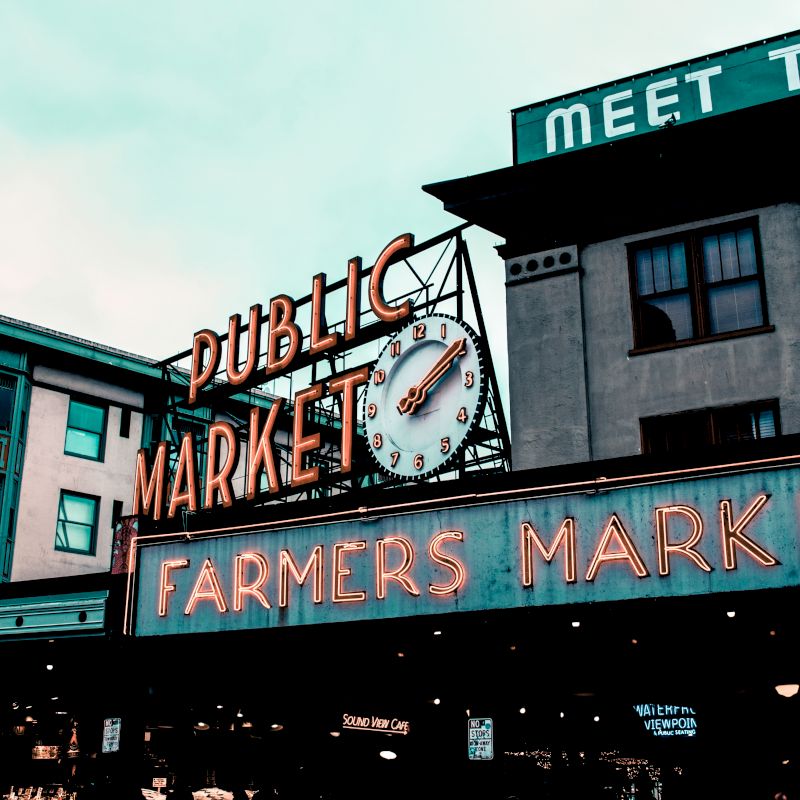 A neon sign reads "Public Market" with a clock above it, and "Farmers Market" below, against a background of buildings.