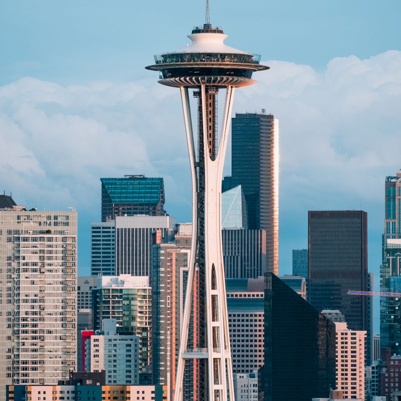 The image shows the Space Needle in Seattle, with a backdrop of the city's skyline featuring various tall buildings and skyscrapers.