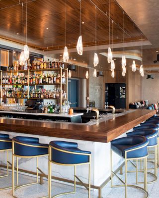 A modern bar with a wooden countertop, blue stools, a liquor display, hanging lights, and tables in the background in a well-lit setting.