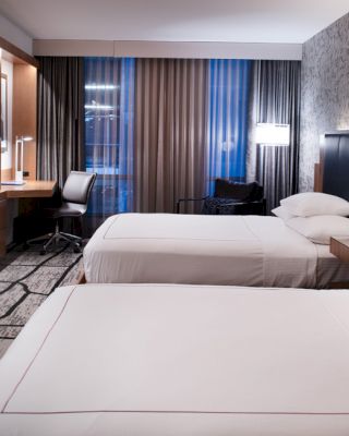 A modern hotel room with two beds, a TV, a desk with a chair, and a well-lit window with curtains.