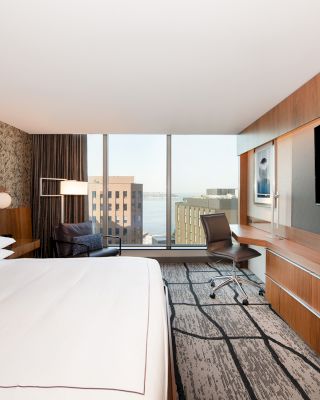 A modern hotel room with a large bed, wall-mounted TV, desk with chair, and a window revealing a city view with buildings and water in the distance.