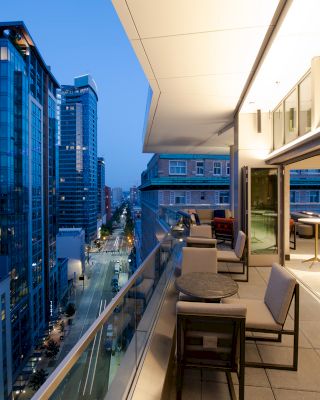 An image features a modern balcony with tables and chairs, overlooking a city street lined with tall buildings at dusk.