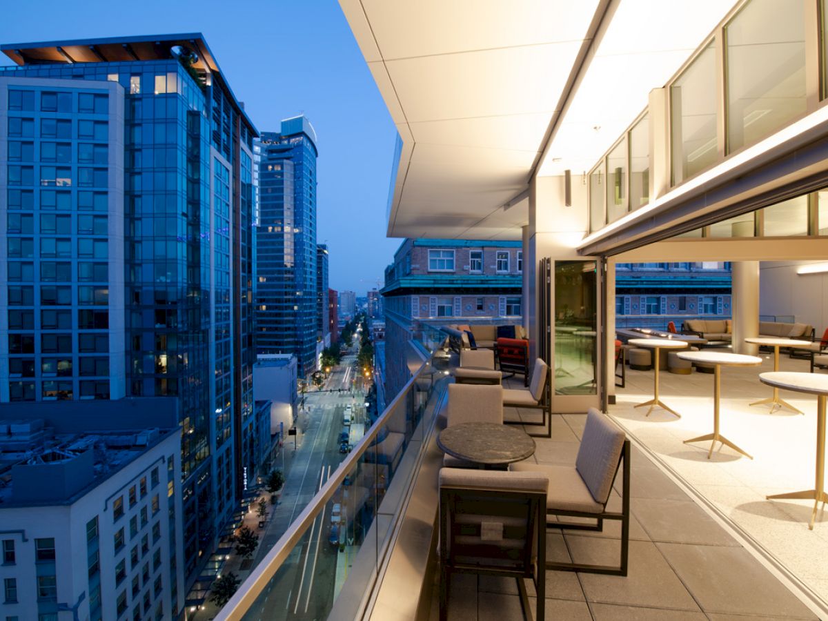 A modern balcony with seating overlooks a cityscape with tall buildings at dusk. Indoor dining area visible through large glass doors.