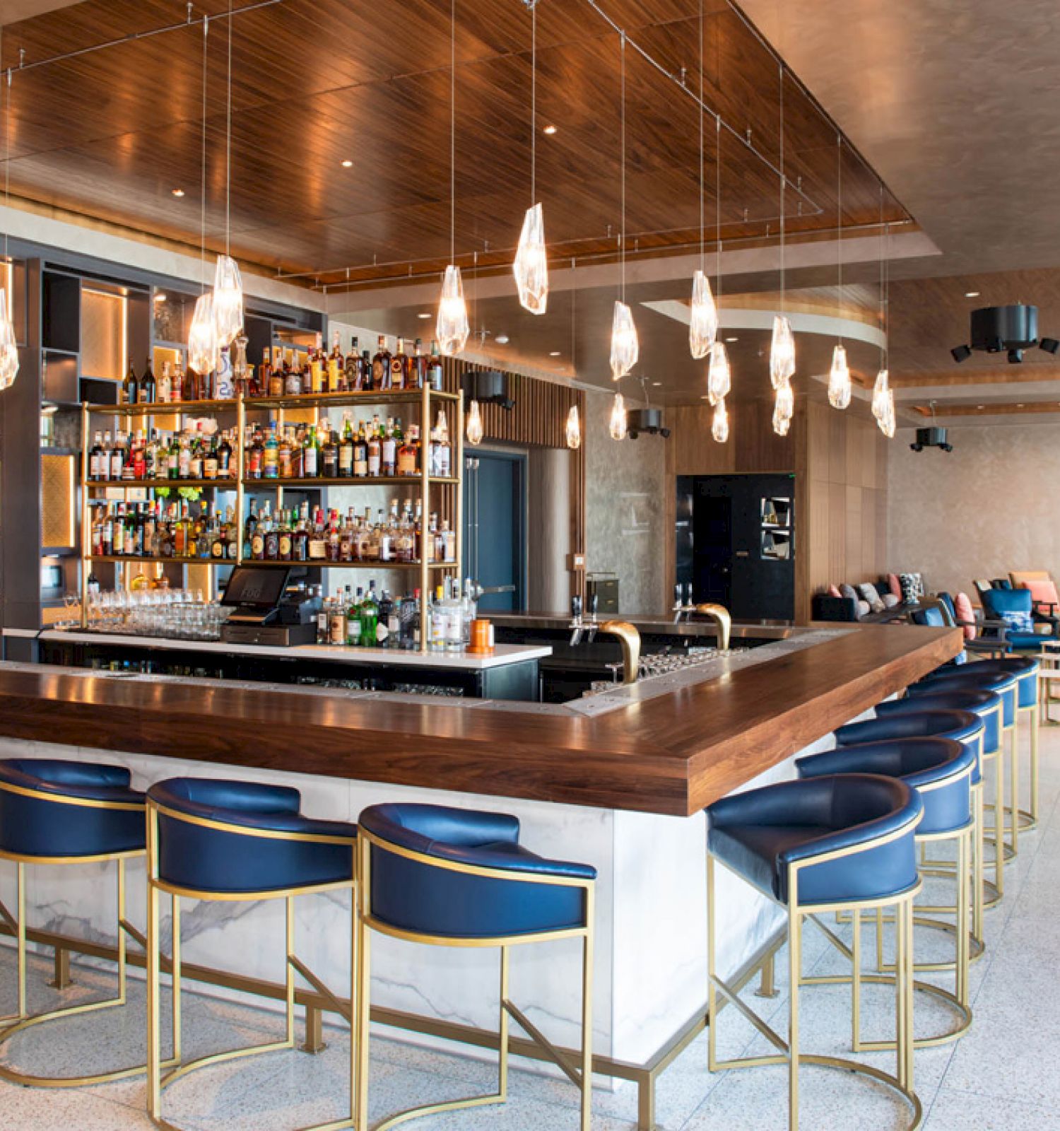 A modern bar with blue stools, a well-stocked liquor shelf, pendant lights, and a seating area in the background with couches and tables.