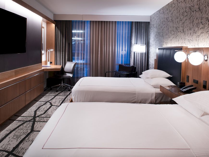 A modern hotel room with two neatly made beds, a wall-mounted TV, a work desk with a chair, and stylish lighting and decor.