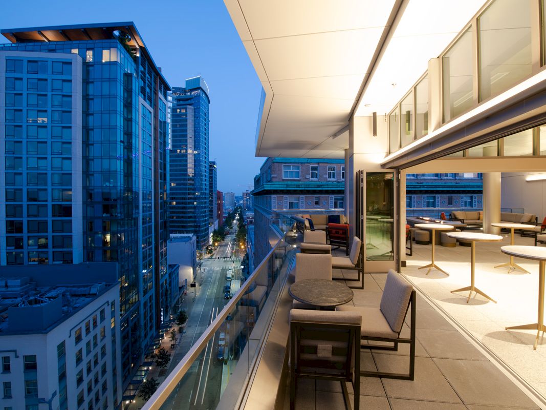 An urban view from a modern balcony overlooking a city street at dusk, featuring outdoor seating and indoor dining areas in the foreground.