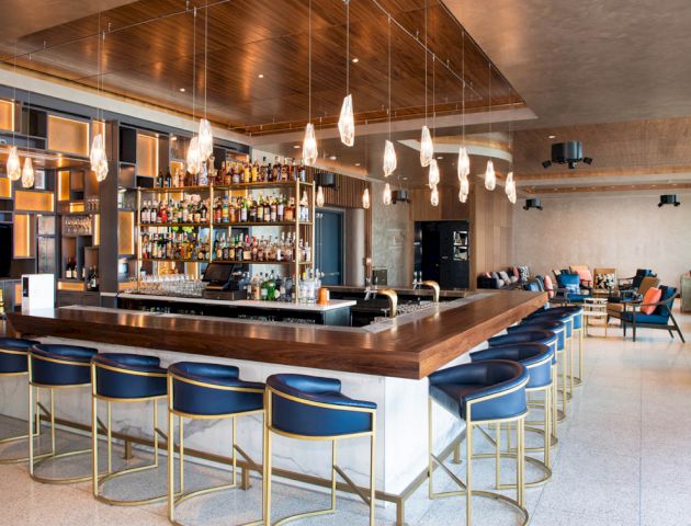 A modern bar with plush seating, overhead lighting, and a well-stocked back bar, featuring blue chairs around a wooden counter.