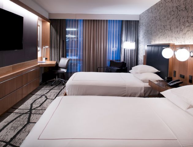 A modern hotel room with two beds, a wall-mounted TV, a desk with a chair, and stylish decor featuring a patterned wall and contemporary lighting.