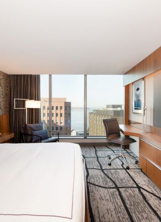 A modern hotel room featuring a large bed, desk with chair, flat-screen TV, and a window with a cityscape and partial water view ends the sentence.
