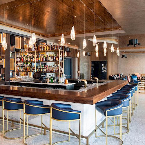 A modern bar with a wooden counter, blue bar stools, hanging lights, and a well-stocked liquor shelf in a stylish, spacious lounge area.