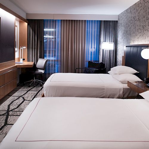 A modern hotel room features two neatly made beds, a wall-mounted TV, a desk with a chair, and large windows with curtains.
