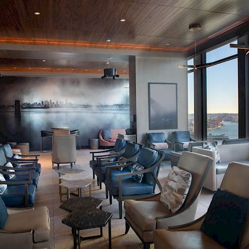 A modern lounge area with comfortable seating, large windows with a scenic view of a waterfront and Ferris wheel, and minimalistic decor.