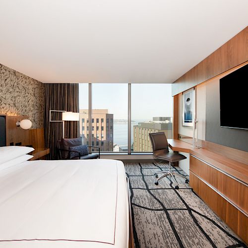 A modern hotel room with a large bed, nightstands, a TV, a desk, and a chair. Large windows provide a view of buildings and a body of water.