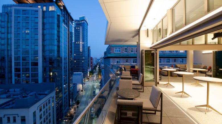A modern rooftop patio with tables and chairs overlooks a city street lined with tall buildings as dusk settles, creating an ambient setting.