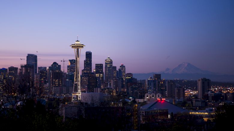 A city skyline at dusk featuring the Space Needle with surrounding buildings and Mount Rainier visible in the background, under a colorful sky.