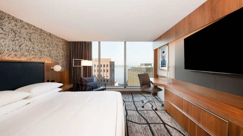 The image shows a modern hotel room with a large bed, wall-mounted TV, desk, chair, and a view of buildings and water through large windows.