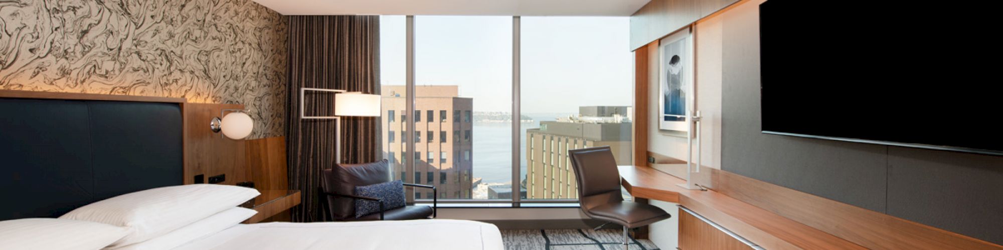 The image shows a modern hotel room with a large bed, a desk and chair, a flat-screen TV, and a view of city buildings through large windows.