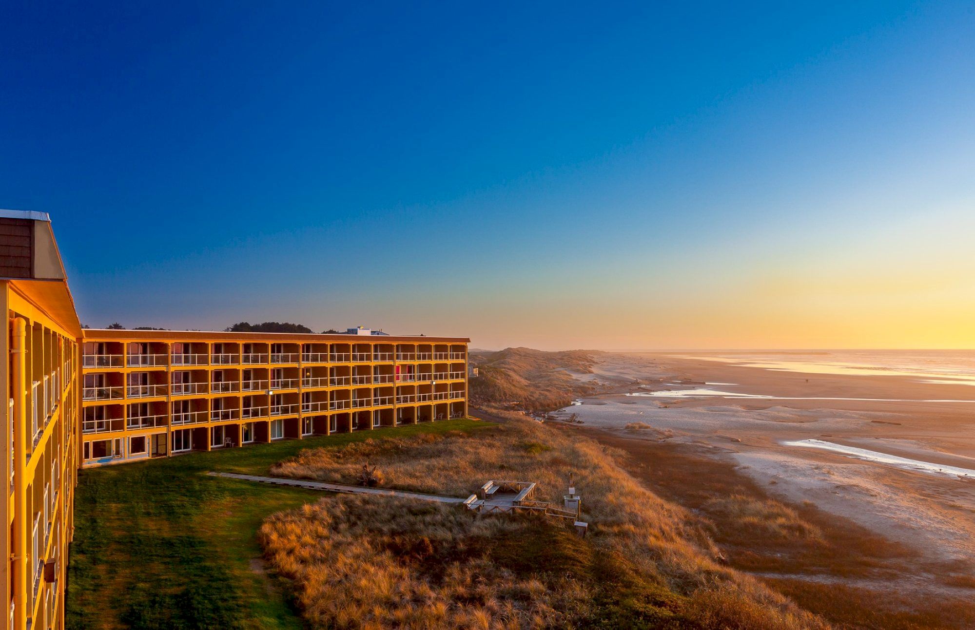 A seaside hotel overlooks a beach at sunset. The sky is clear and the landscape is serene, with grassy dunes leading to the shore.